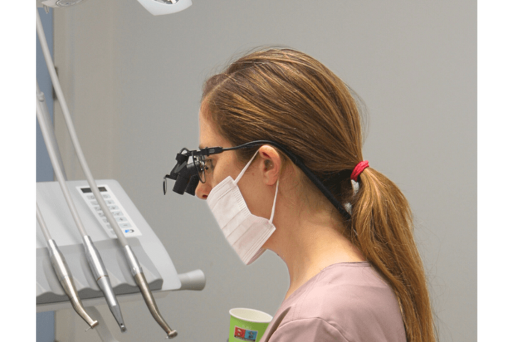 A woman in a dental office wearing magnifying glasses, a white face mask, and a pink scrub top, shown in profile near dental equipment.