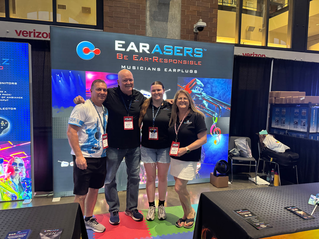 Four people posing together in front of a large 'EARASERS Be Ear-Responsible Musicians Earplugs' booth at a convention.