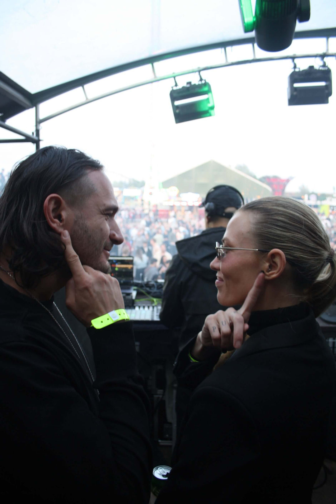 a man and woman look at each other smiling on stage in front of a crowd while pointing to the earplugs in their ears.