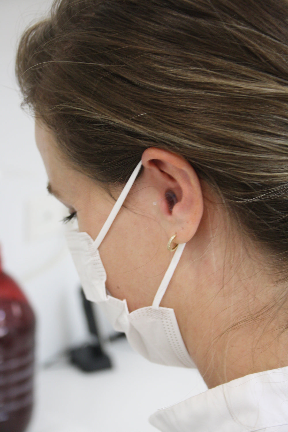 A close-up side profile of a woman hygienist wearing a white face mask and a small gold hoop earring, with her hair and ear clearly visible against a softly blurred indoor background.  She is wearing earasers.