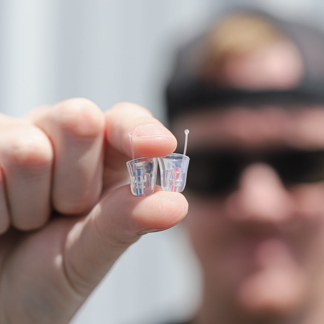 A man in sunglasses holds up a pair of clear Earasers between his fingers