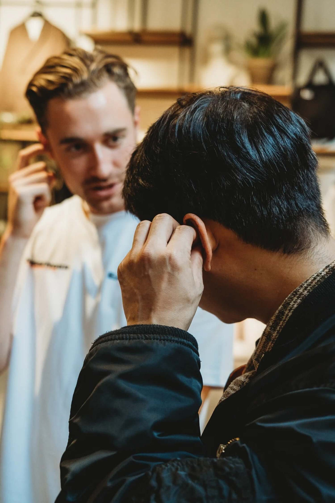 A man in a black jacket adjusts one of his Earasers in his ear while another man in a white shirt looks on in a softly lit indoor setting.
