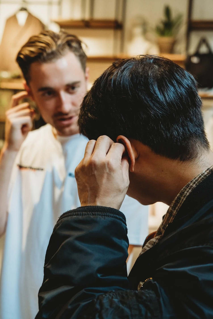 A man in a black jacket adjusts one of his Earasers in his ear while another man in a white shirt looks on in a softly lit indoor setting.
